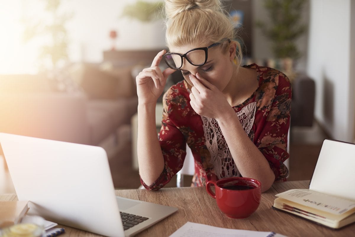 Mulher de óculos e blusa estampada, sentada diante de um laptop, com a mão no rosto em expressão de cansaço, uma caneca vermelha sobre a mesa de madeira