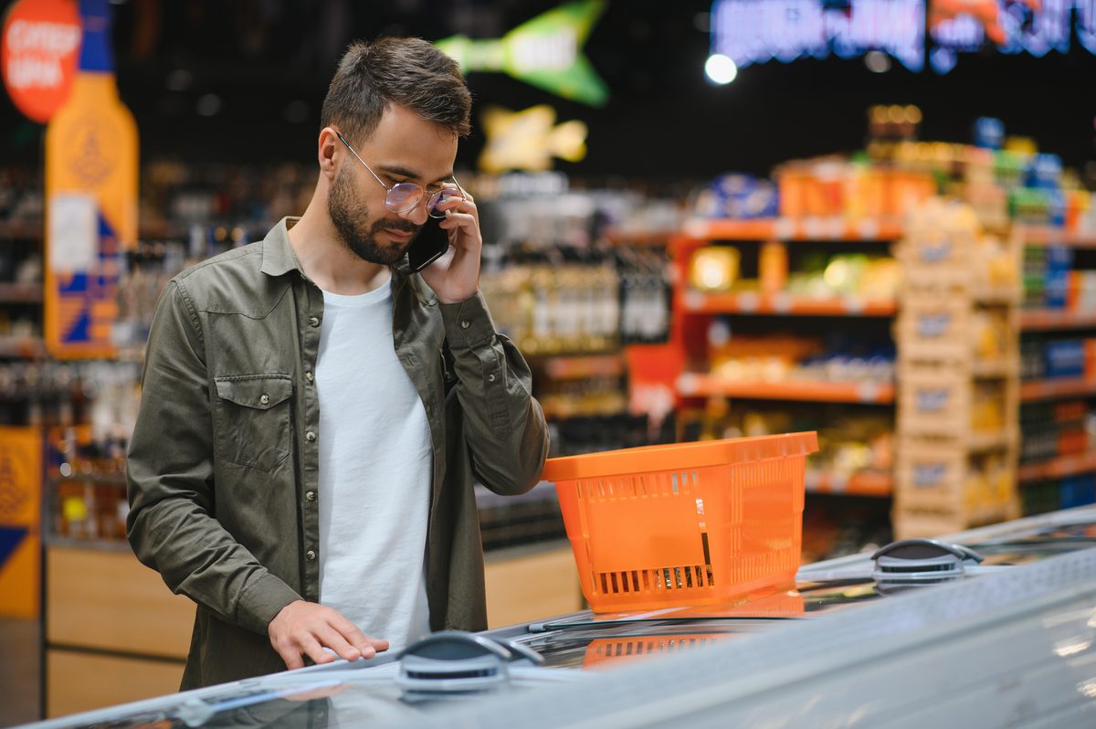 Homem de camisa verde e óculos fala ao celular enquanto observa produtos em um freezer aberto de supermercado, com uma cesta de compras laranja apoiada na borda