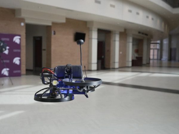 Pequeno drone de hélices protegidas paira no corredor de uma escola, com piso claro, colunas de tijolo e um cartaz roxo ao fundo