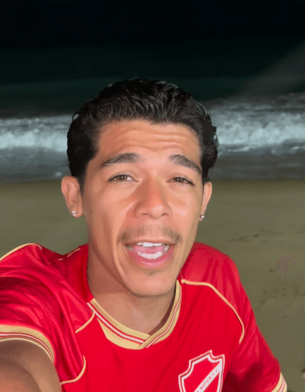 Jovem sorridente em selfie na praia, usando camisa esportiva vermelha