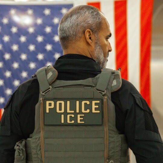 Agente com colete tático escrito ICE Police visto de costas, com bandeira dos Estados Unidos ao fundo