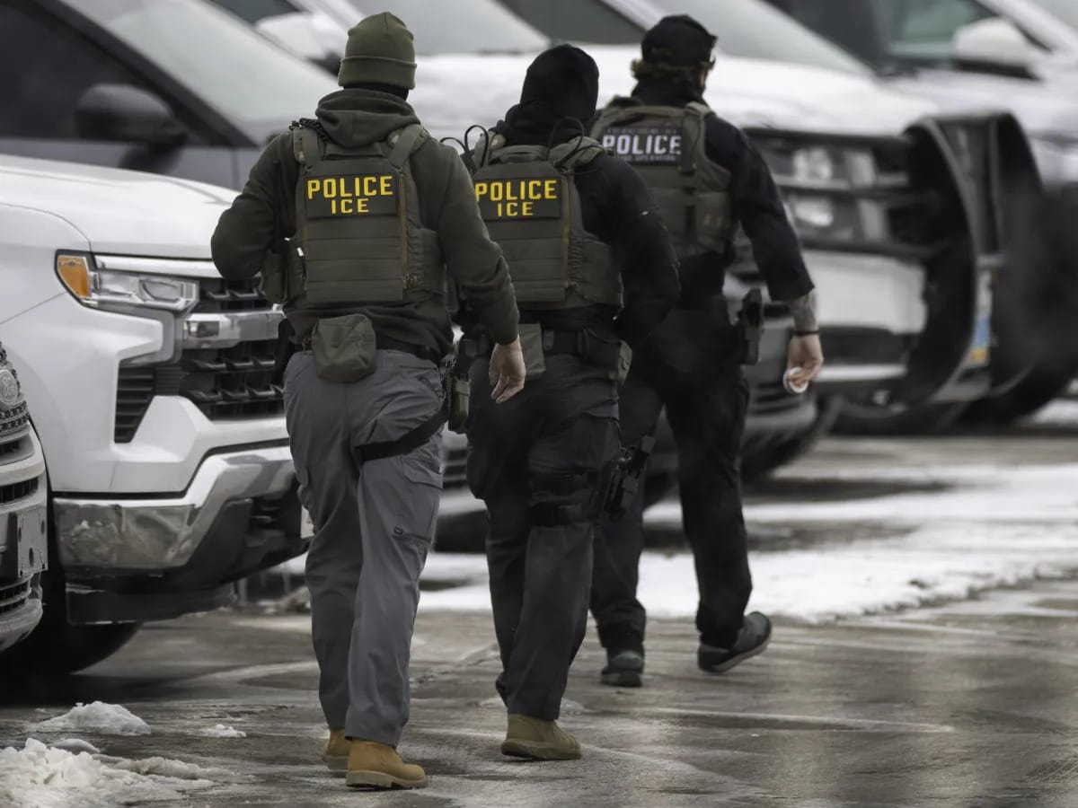 Três agentes com colete escrito Police ICE caminhando entre viaturas policiais