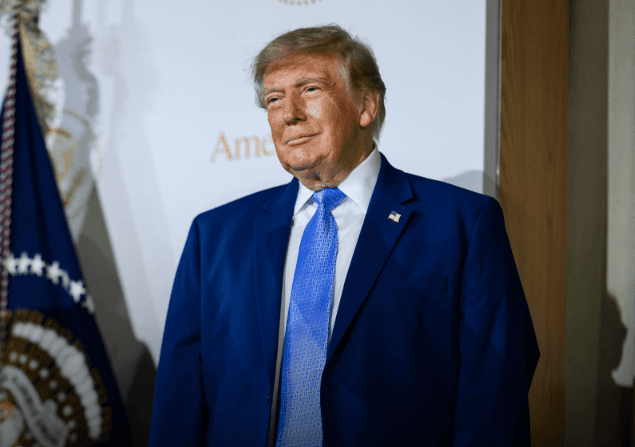 Donald Trump, de terno azul, ao lado da bandeira presidencial dos Estados Unidos