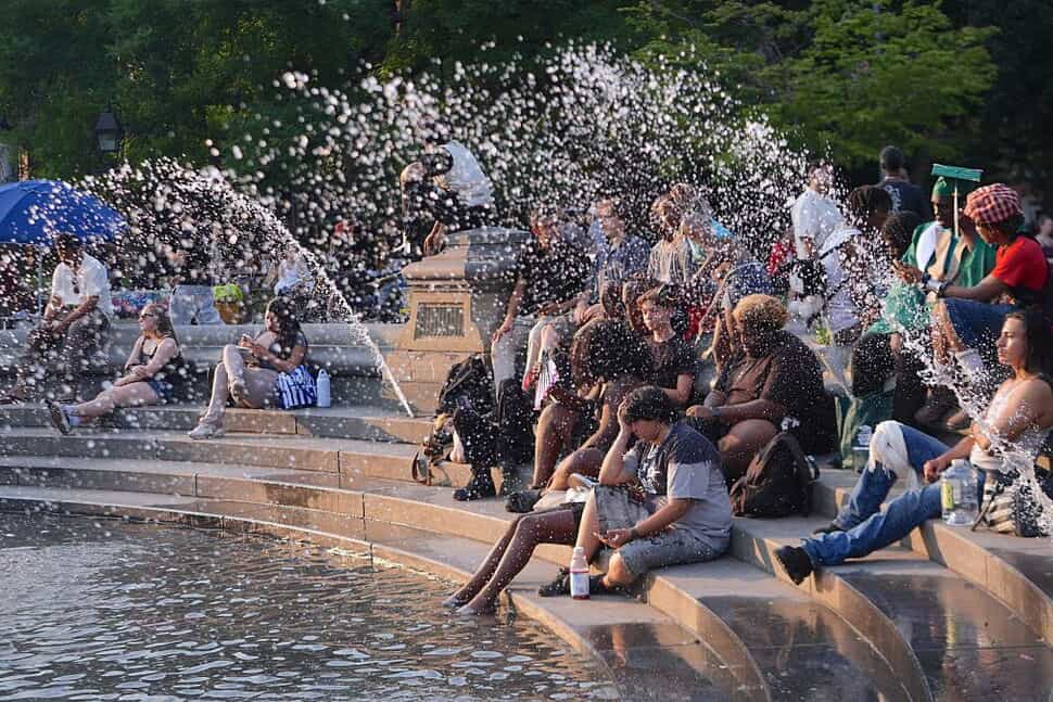 Pessoas sentadas na borda de uma fonte pública se refrescam com os jatos d'água em dia de calor extremo