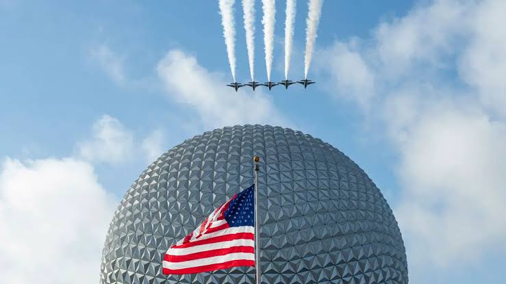 Cinco aeronaves militares em formação deixam trilhas de fumaça sobre a esfera do EPCOT, com a bandeira dos EUA em primeiro plano