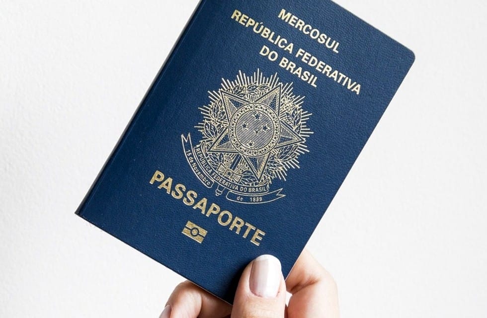 Mão segura um passaporte brasileiro azul, com o brasão dourado da República Federativa do Brasil e a inscrição Mercosul na capa