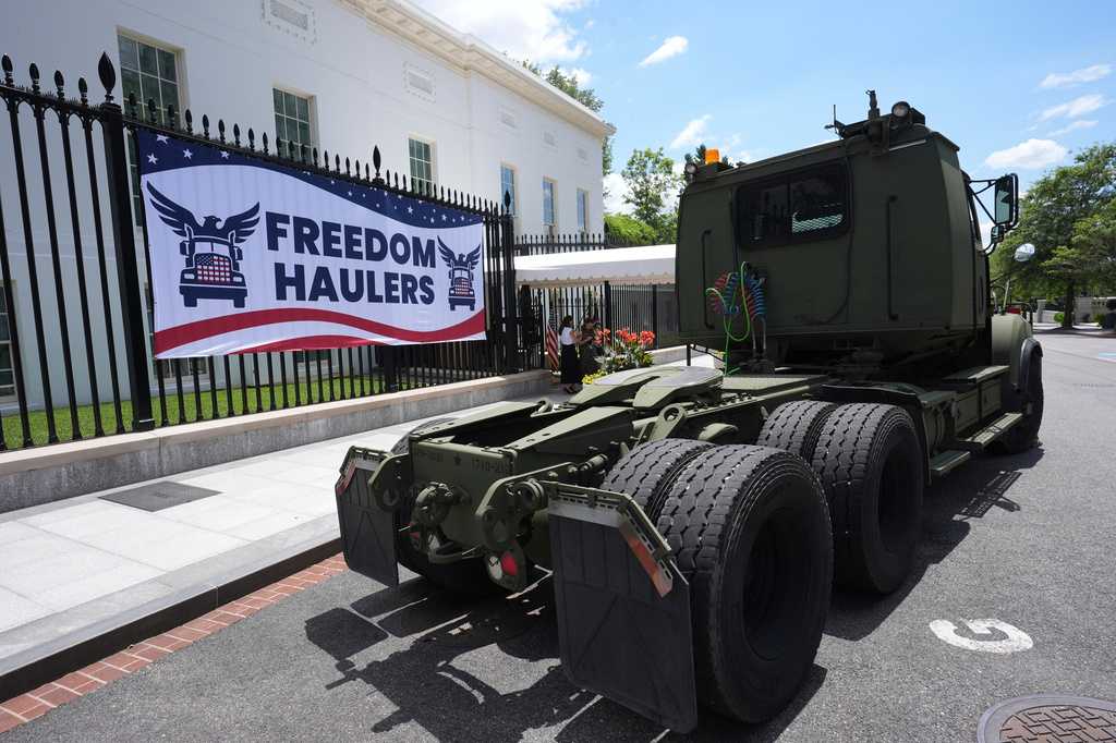 Caminhão militar verde estacionado diante da grade da Casa Branca, ao lado de um banner vermelho, branco e azul com o logotipo do programa Freedom Haulers