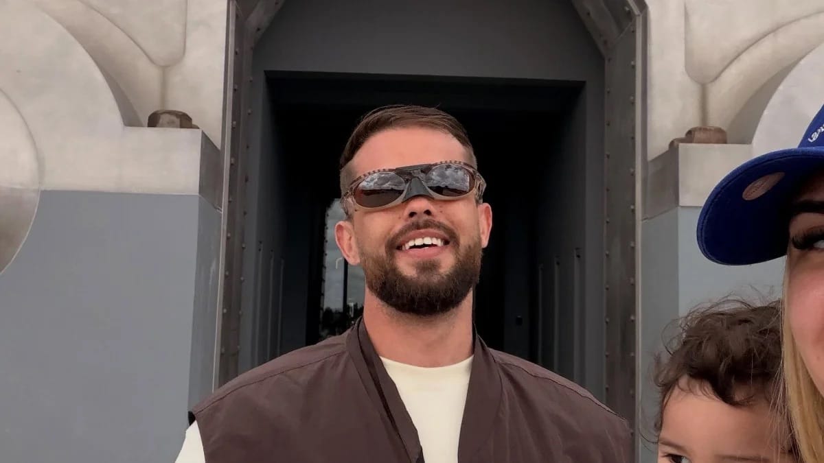 Homem jovem de barba, óculos escuros na testa e colete marrom sorri em foto de família, ao lado de uma mulher de boné azul e uma criança pequena