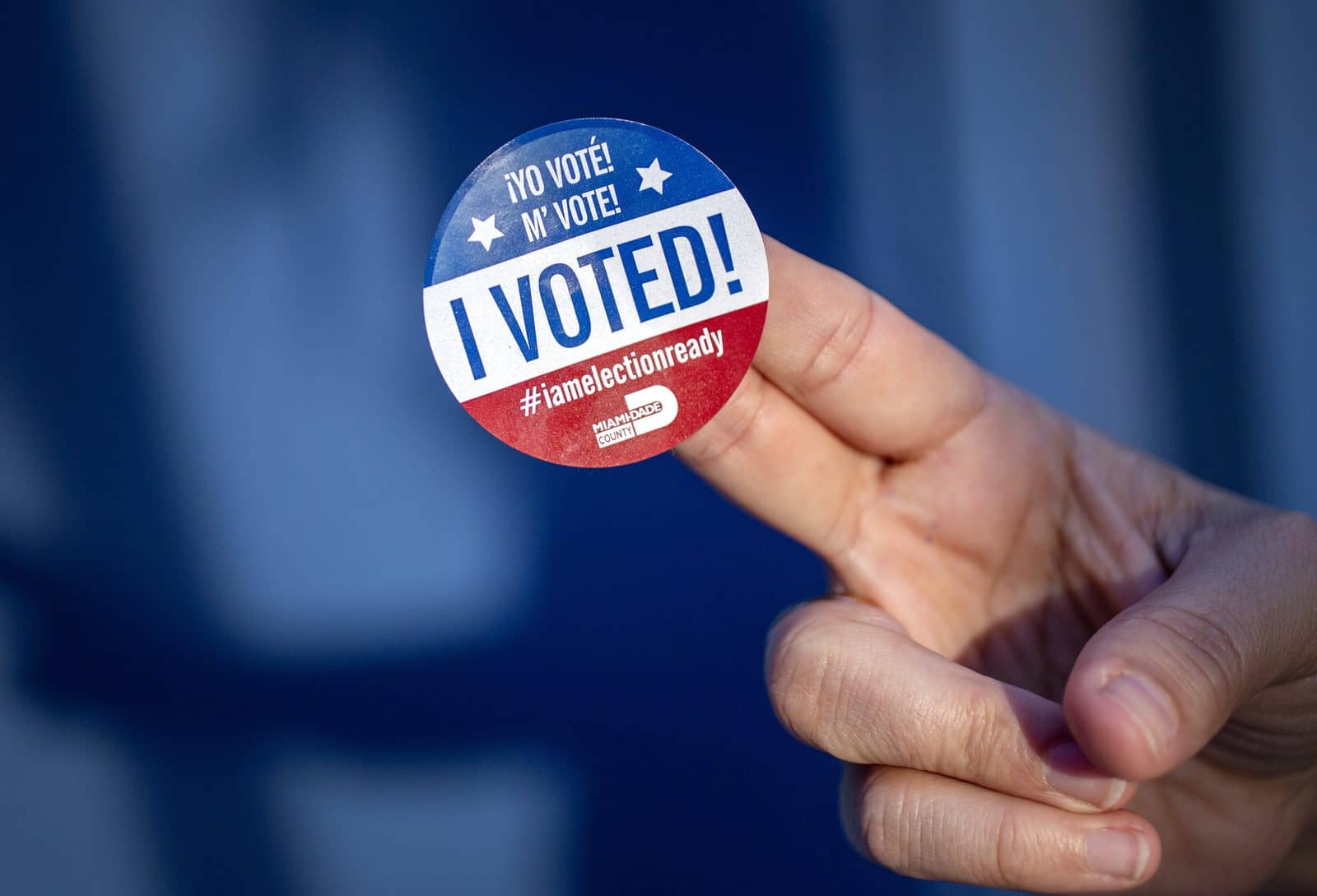 Mão segura um adesivo redondo de urna com as frases I Voted, Yo Voté e M' Vote e o logotipo do condado de Miami-Dade, sobre fundo azul desfocado