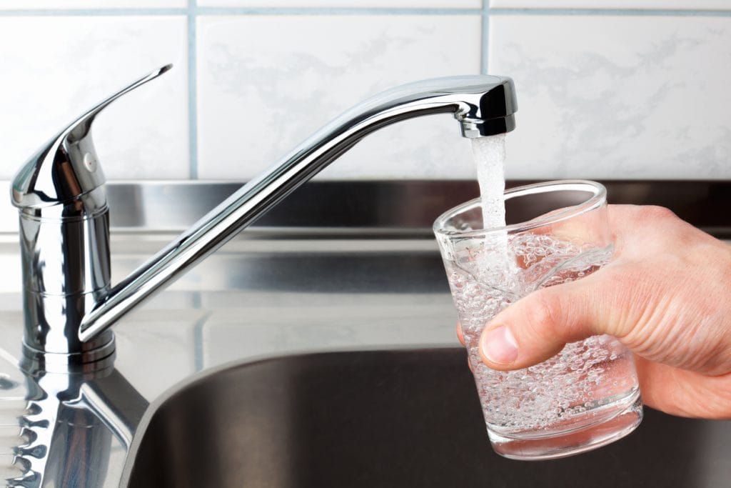 Mão segura um copo de vidro que é enchido com água corrente na torneira de uma pia de cozinha em aço inox