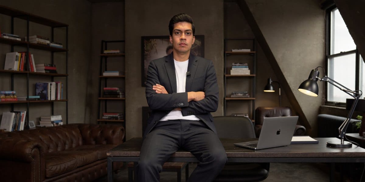 Darley Tomaz, de blazer escuro e camiseta branca, sentado na borda de uma mesa de escritório com braços cruzados, entre estantes de livros e um notebook