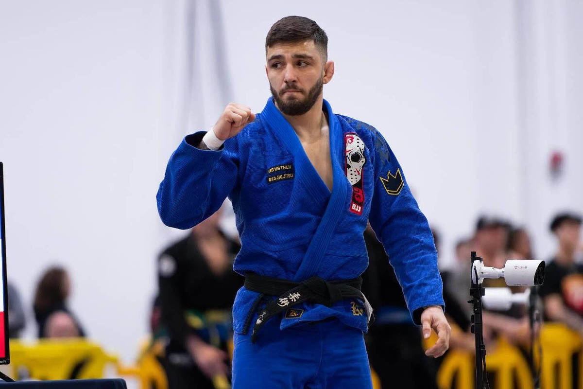 Homem jovem de barba, quimono azul e faixa-preta de jiu-jítsu, com o punho erguido, durante competição em ginásio