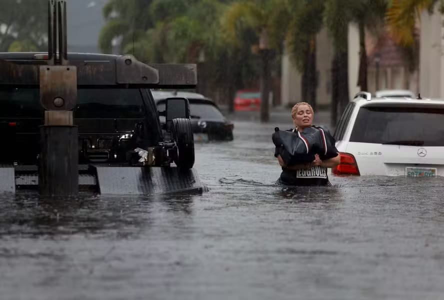 Mulher atravessa rua alagada com água na altura da cintura, carregando uma bolsa acima da água, entre carros parcialmente submersos sob chuva forte