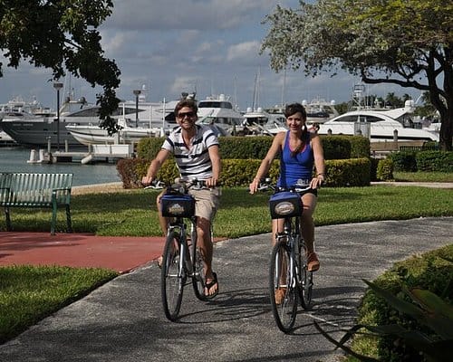 Duas pessoas pedalando bicicletas em calçada à beira de uma marina, com barcos ao fundo