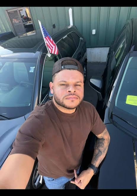 Homem jovem de barba curta, boné preto virado para trás e camiseta marrom, em selfie ao lado de carros, com uma bandeira dos Estados Unidos ao fundo