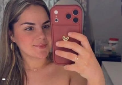Mulher jovem de cabelos castanhos claros, brincos de argola e colar fino, em selfie diante do espelho segurando um celular com capa rosa