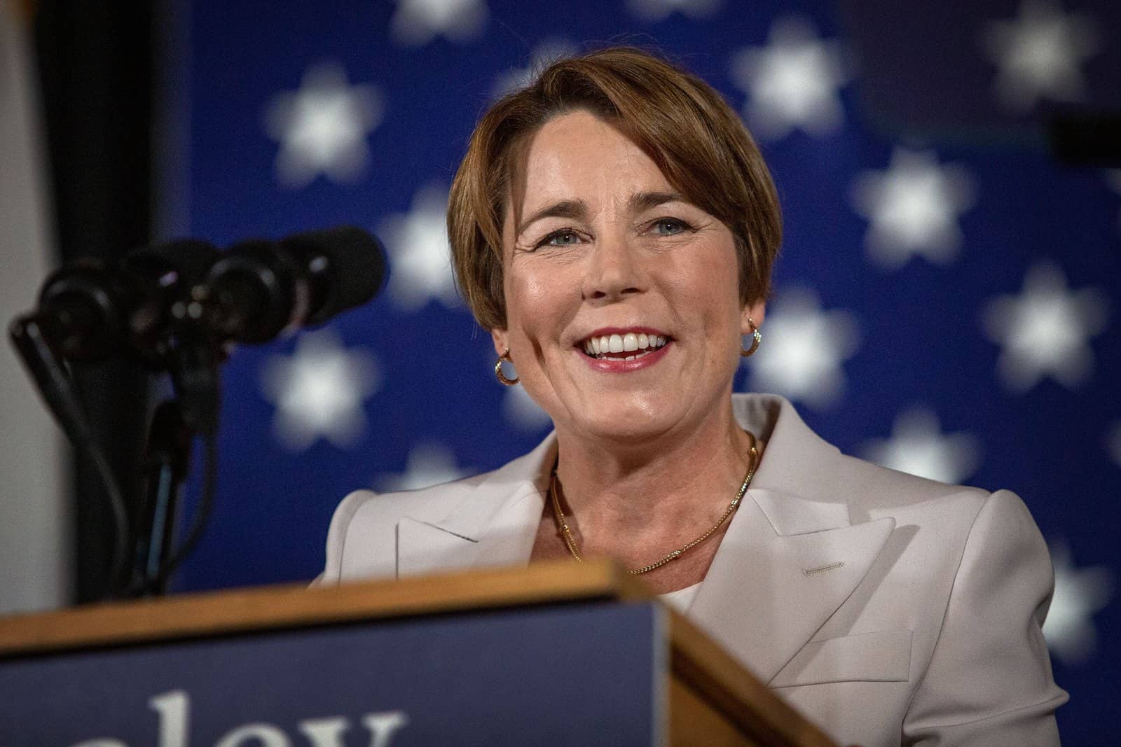 Governadora Maura Healey sorrindo atrás de um púlpito com microfone, diante de um painel azul com estrelas brancas