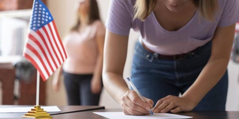 Mulher preenchendo um formulário sobre uma mesa com caneta azul, ao lado de uma pequena bandeira dos Estados Unidos em base dourada