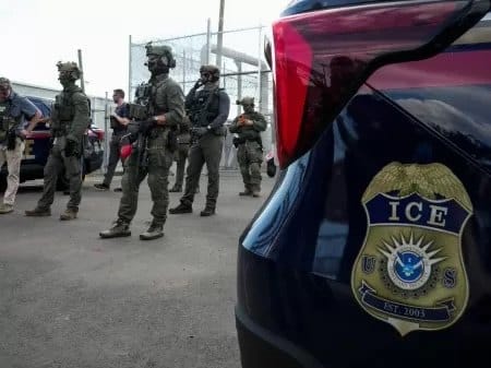 Agentes federais com equipamento tático em pé diante de uma cerca, ao lado de veículo com o brasão do ICE em destaque