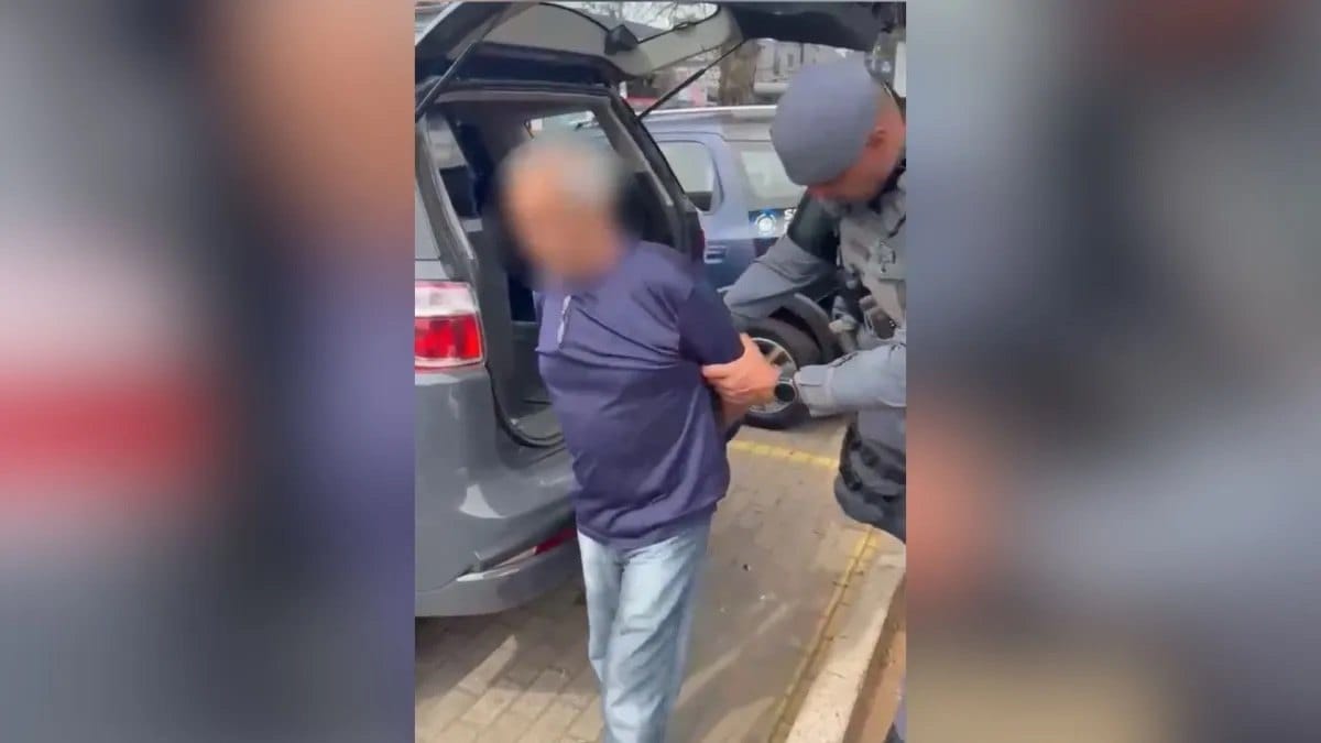 Policial fardado conduz homem de camiseta escura, com o rosto borrado, ao lado do porta-malas aberto de um carro em via pública