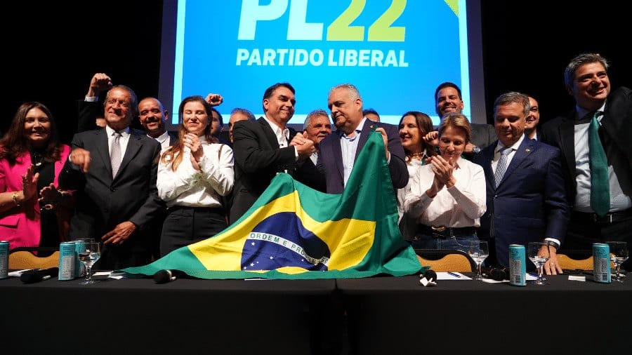 Flávio Bolsonaro e Alfredo Gaspar de mãos dadas erguidas atrás de uma bandeira do Brasil, cercados por parlamentares que aplaudem, com painel do PL ao fundo