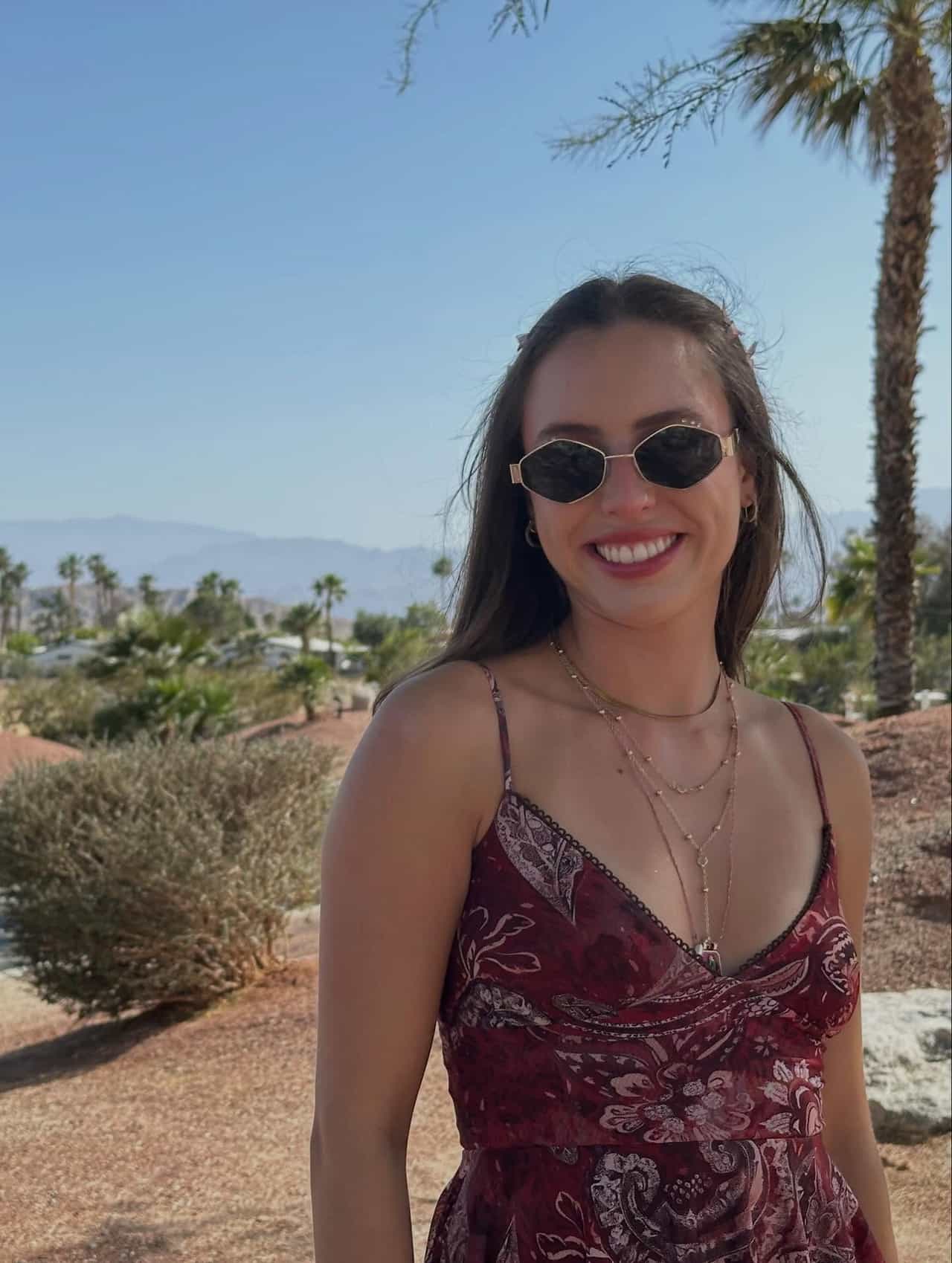Mulher sorrindo, de óculos escuros e vestido estampado vermelho, em paisagem desértica com palmeiras