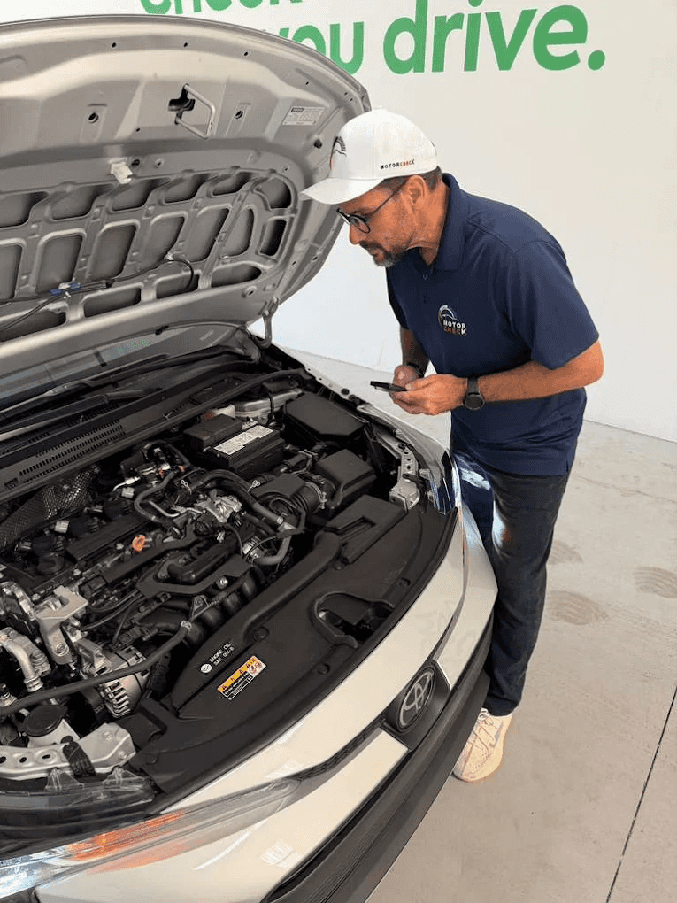 Técnico da MotorCheck, de boné e camisa da marca, inspeciona o motor de um carro com o capô aberto, anotando dados no celular