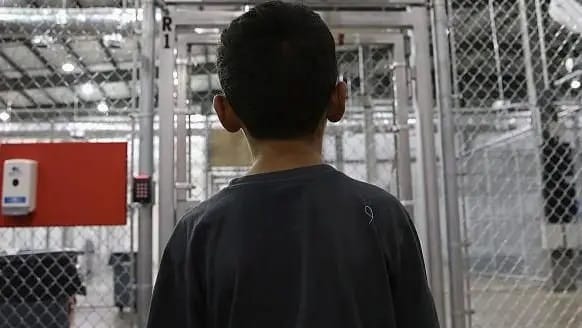 Menino de costas, de camiseta cinza escura, em pé dentro de instalação com divisórias de tela metálica e luzes de galpão ao fundo