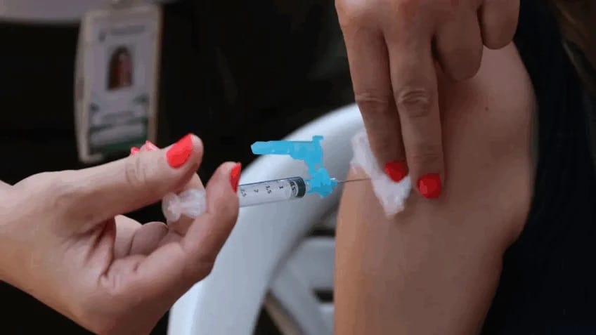 Profissional de saúde aplica injeção no braço de uma paciente sentada, segurando algodão com a outra mão