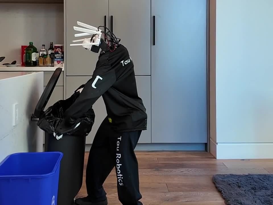 Robô humanoide vestindo agasalho preto com a marca Tau Robotics troca o saco de uma lixeira em cozinha de apartamento, ao lado de um cesto de reciclagem azul