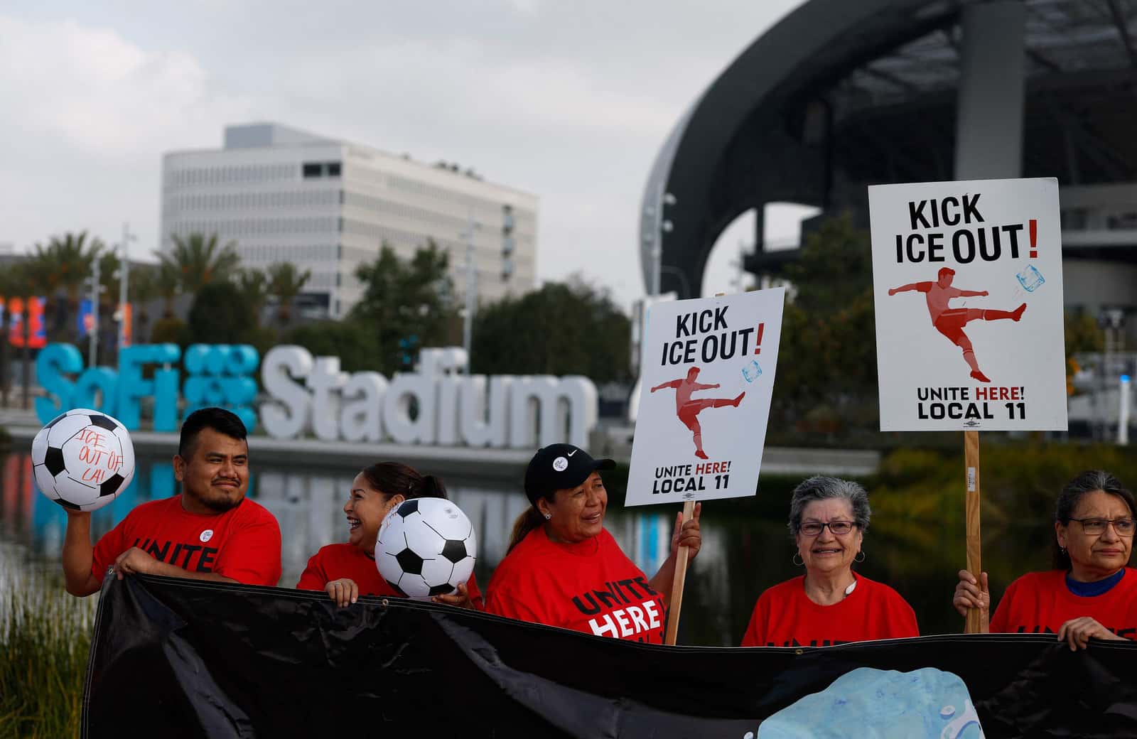 Manifestantes de camiseta vermelha do sindicato UNITE HERE Local 11 erguem cartazes com os dizeres 'Kick ICE Out' em frente ao SoFi Stadium, em Los Angeles