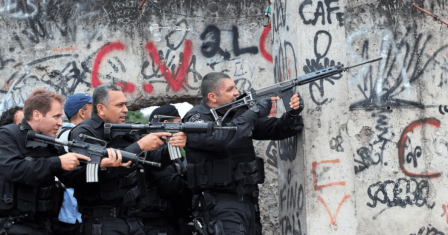 Policiais fortemente armados, com fuzis e coletes táticos, se posicionam atrás de um muro de concreto pichado em uma comunidade no Brasil
