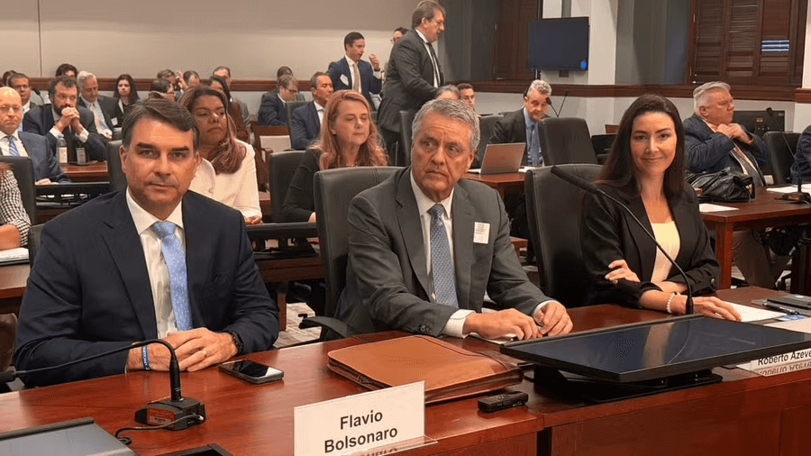 O senador Flávio Bolsonaro sentado a uma mesa de audiência com uma placa com seu nome, ao lado de outros participantes, em sala cheia de público de terno