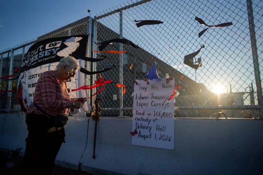 Mulher de camisa xadrez amarra fitas vermelhas no alambrado de um centro de detenção, ao lado de um cartaz manuscrito com o nome de Edwin Jovanny Lopez Cornejo e a data de sua morte