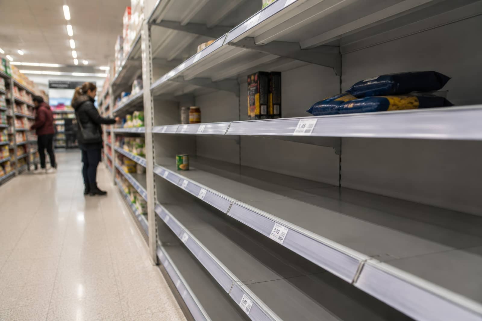 Corredor de supermercado com prateleiras vazias e duas pessoas ao fundo