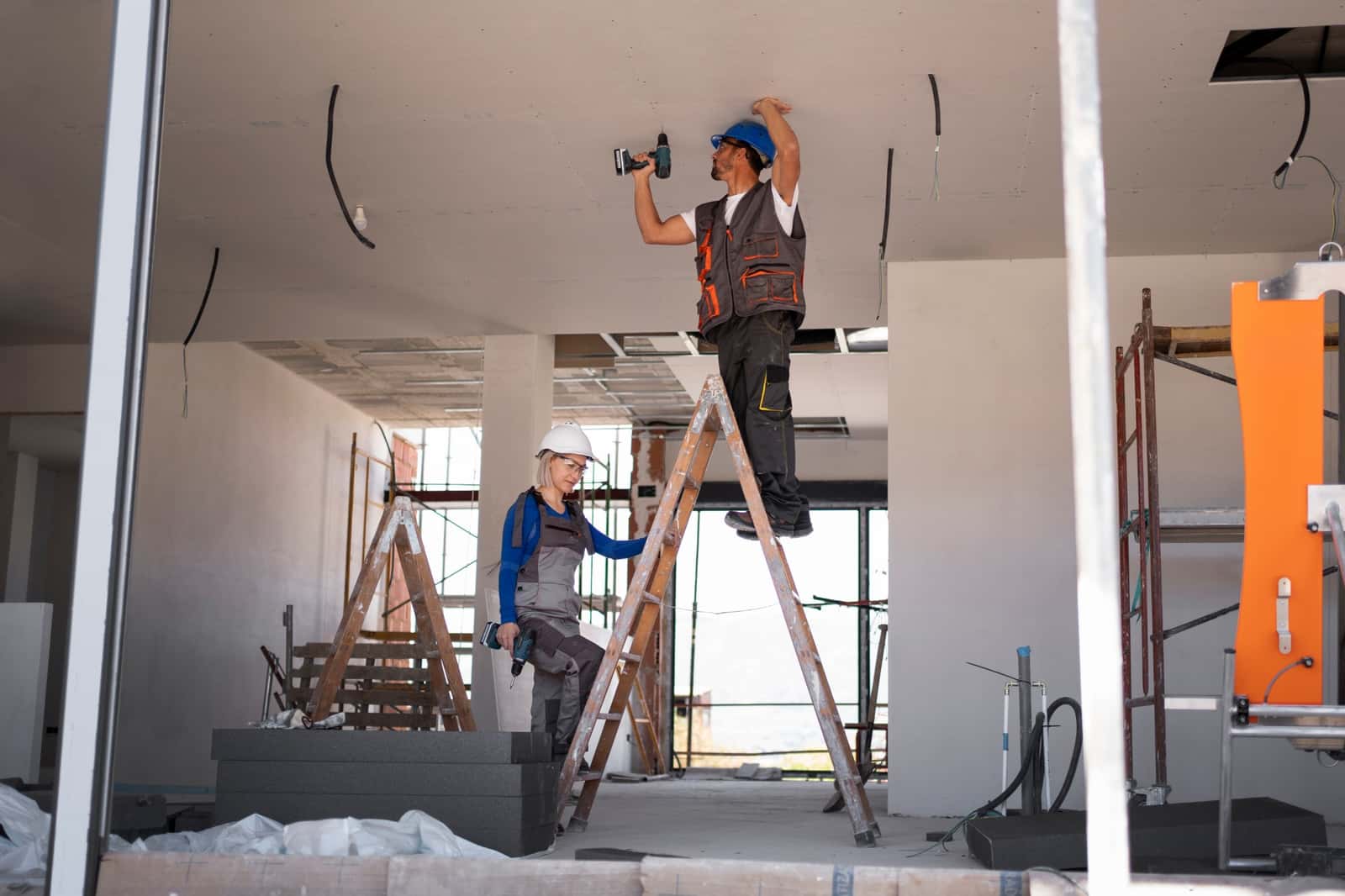 Dois trabalhadores de capacete em obra, um deles em uma escada, furando o teto