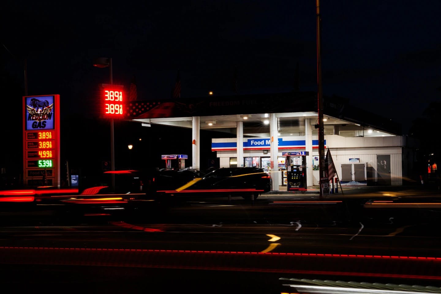 Posto de gasolina iluminado à noite com o letreiro Freedom Fuel na marquise, painel de preços em vermelho marcando 3,89 e rastros de faróis de carros na avenida em frente
