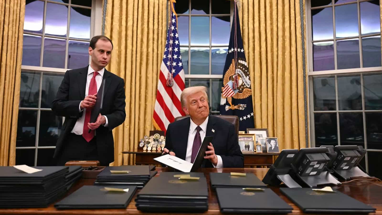 Presidente dos Estados Unidos sentado à mesa do Salão Oval segura uma pasta de decreto assinada, com pilhas de pastas pretas à frente e um assessor em pé ao lado