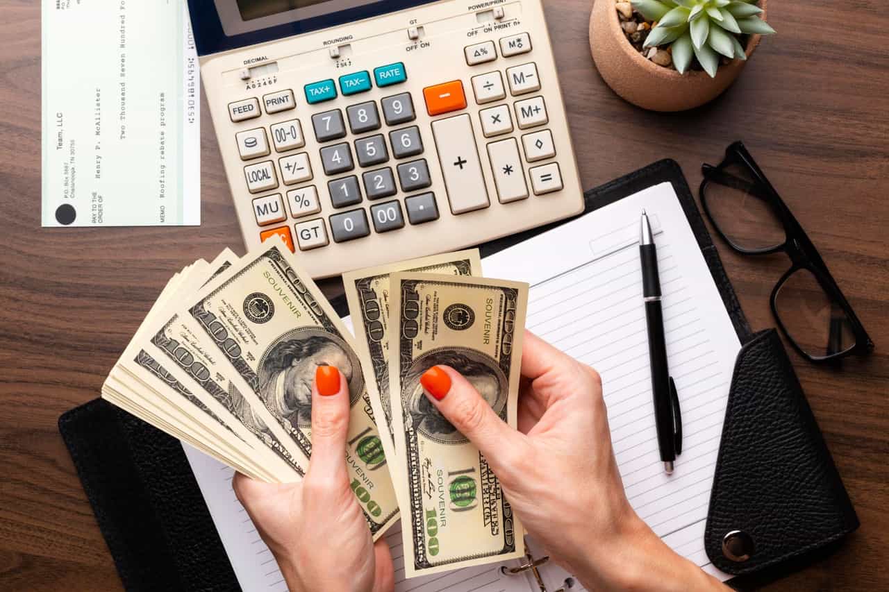 Mãos contando notas de 100 dólares sobre mesa com calculadora, caderno, óculos e um cheque
