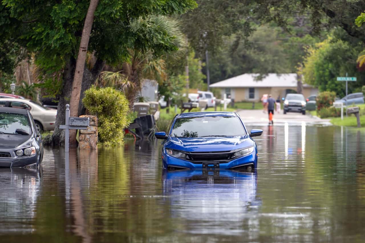 Rua residencial alagada, com um carro azul parcialmente submerso e outro veículo ao lado, e pessoas caminhando ao fundo