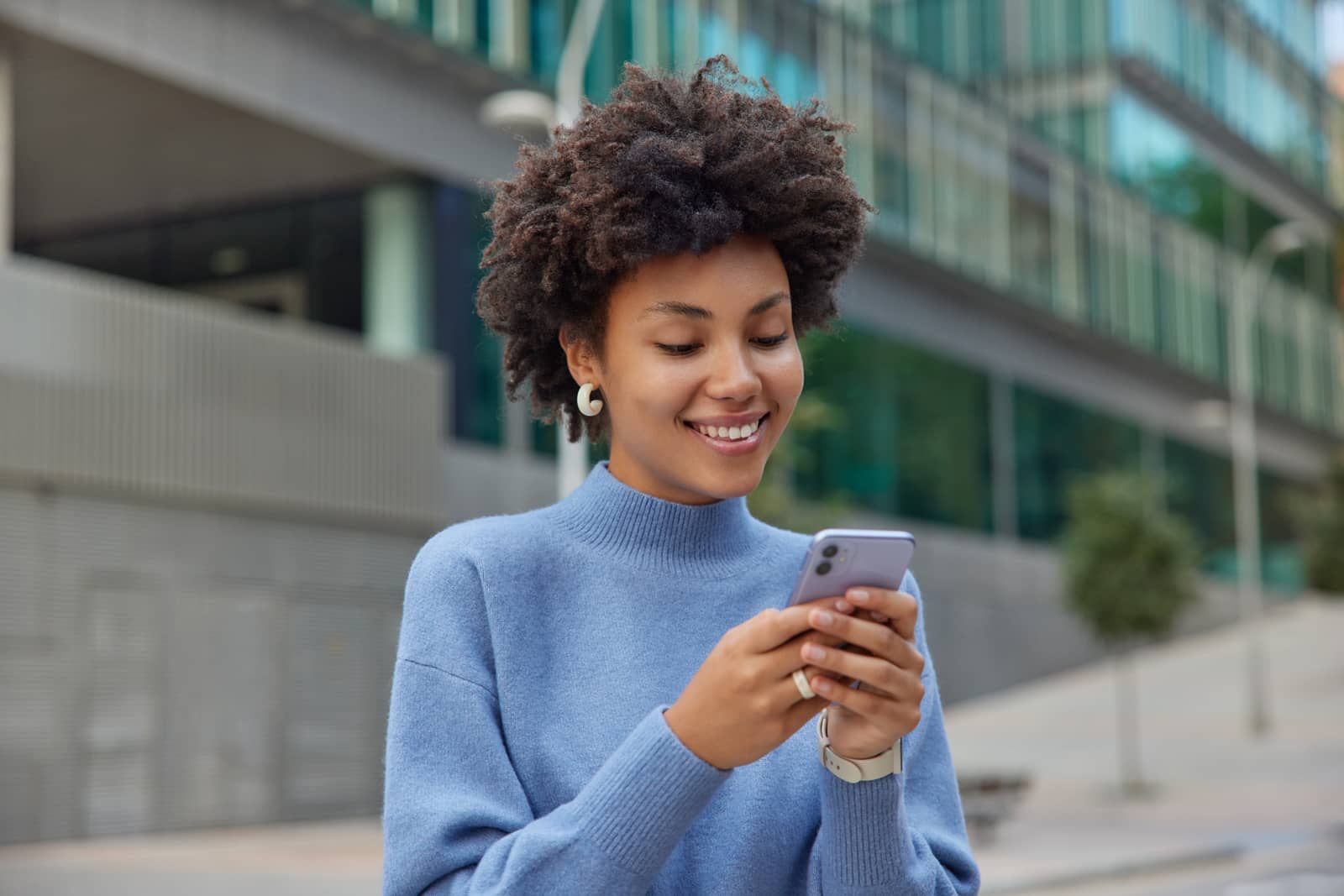 Mulher sorri ao olhar o celular em uma rua com prédios de vidro ao fundo
