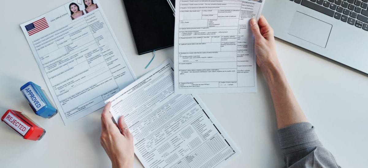 Vista de cima de mãos preenchendo e comparando formulários de visto americano sobre uma mesa, com carimbos de "approved" e "rejected" ao lado e um notebook ao fundo