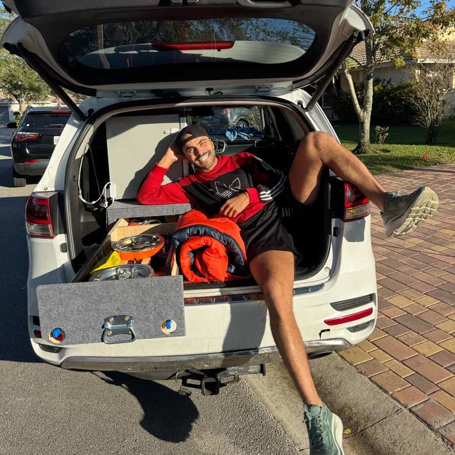 Bruno Freitas sorrindo deitado no porta-malas do carro adaptado, com fogareiro portátil e saco de dormir