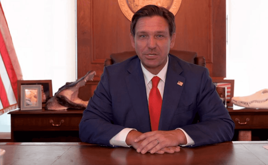 Governador da Flórida, Ron DeSantis, de terno azul-marinho e gravata vermelha, sentado à mesa de trabalho com o selo do estado ao fundo e cabeça de jacaré empalhada sobre um móvel lateral