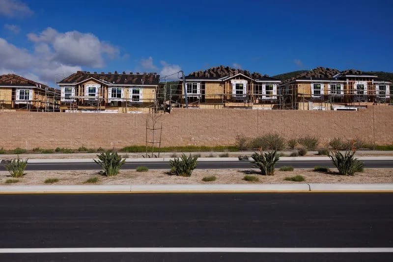 Fileira de casas em construção, com estrutura de madeira aparente e andaimes, atrás de um muro bege, vistas do outro lado de uma avenida recém-pavimentada