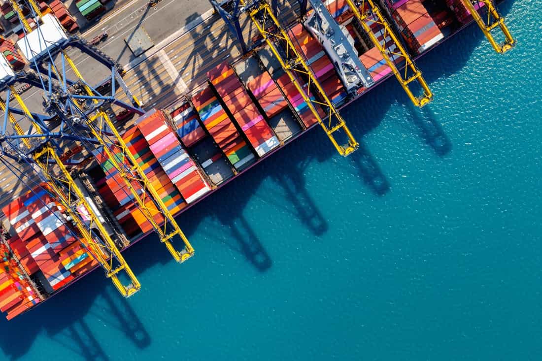 Vista aérea de navio de carga atracado em porto, com contêineres coloridos, guindastes amarelos e mar azul