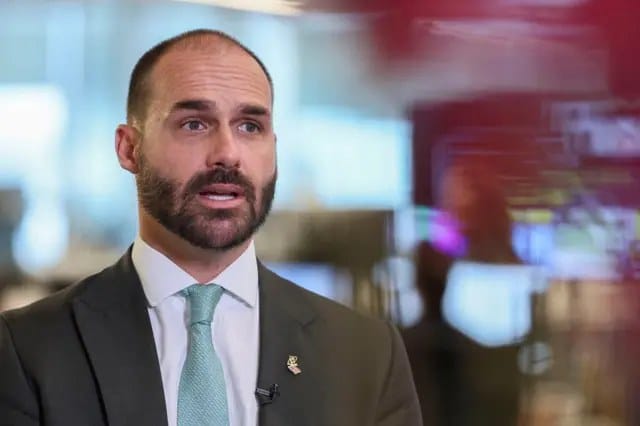 Homem de barba escura, de terno e gravata verde-clara, fala em uma entrevista, com um microfone de lapela