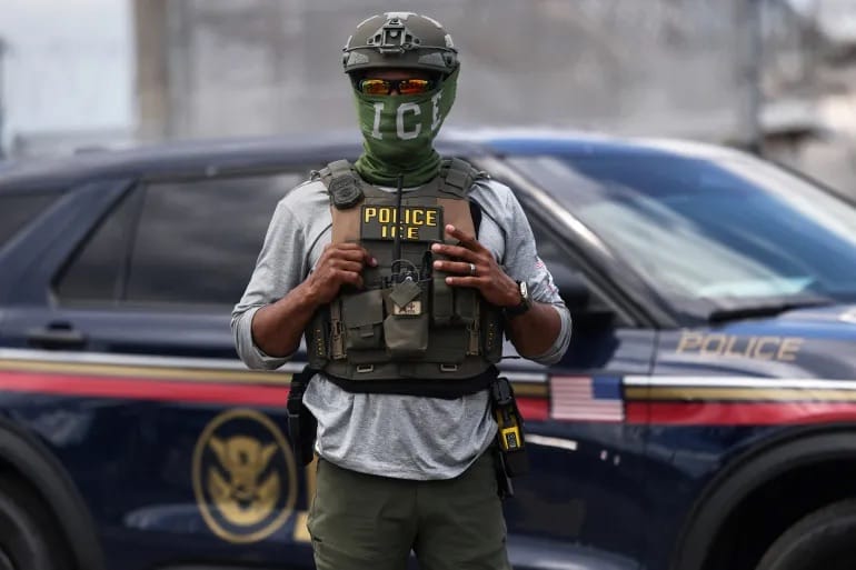 Agente do ICE com o rosto coberto por uma bandana com a inscrição ICE e colete tático escrito Police ICE, diante de uma viatura escura