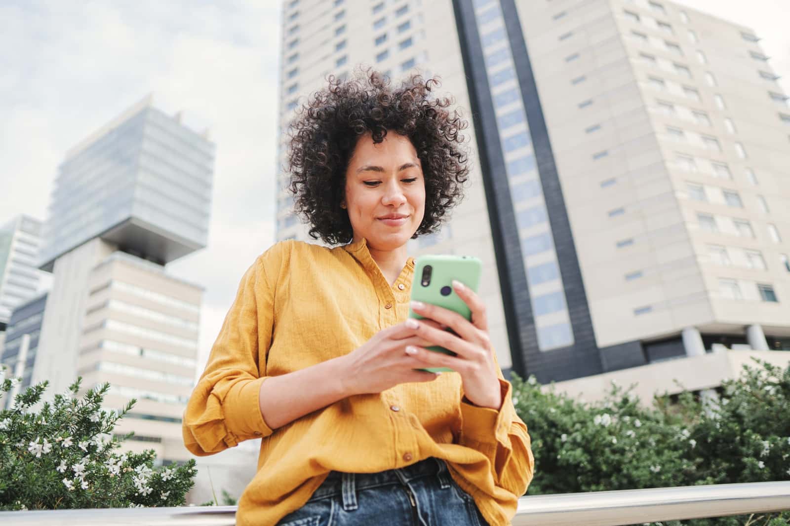 Mulher de camisa amarela sorri ao usar um aplicativo no smartphone em uma rua, com prédios altos ao fundo