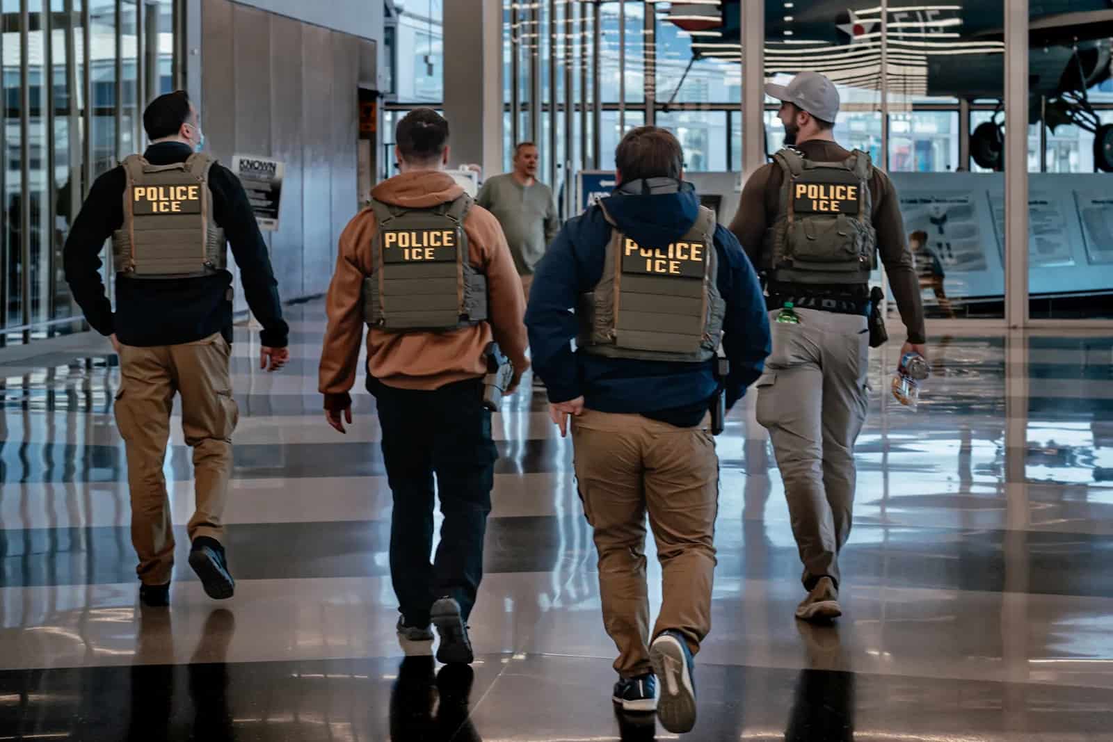 Quatro agentes vistos de costas caminham por um saguão envidraçado usando coletes táticos com a inscrição POLICE ICE em letras amarelas